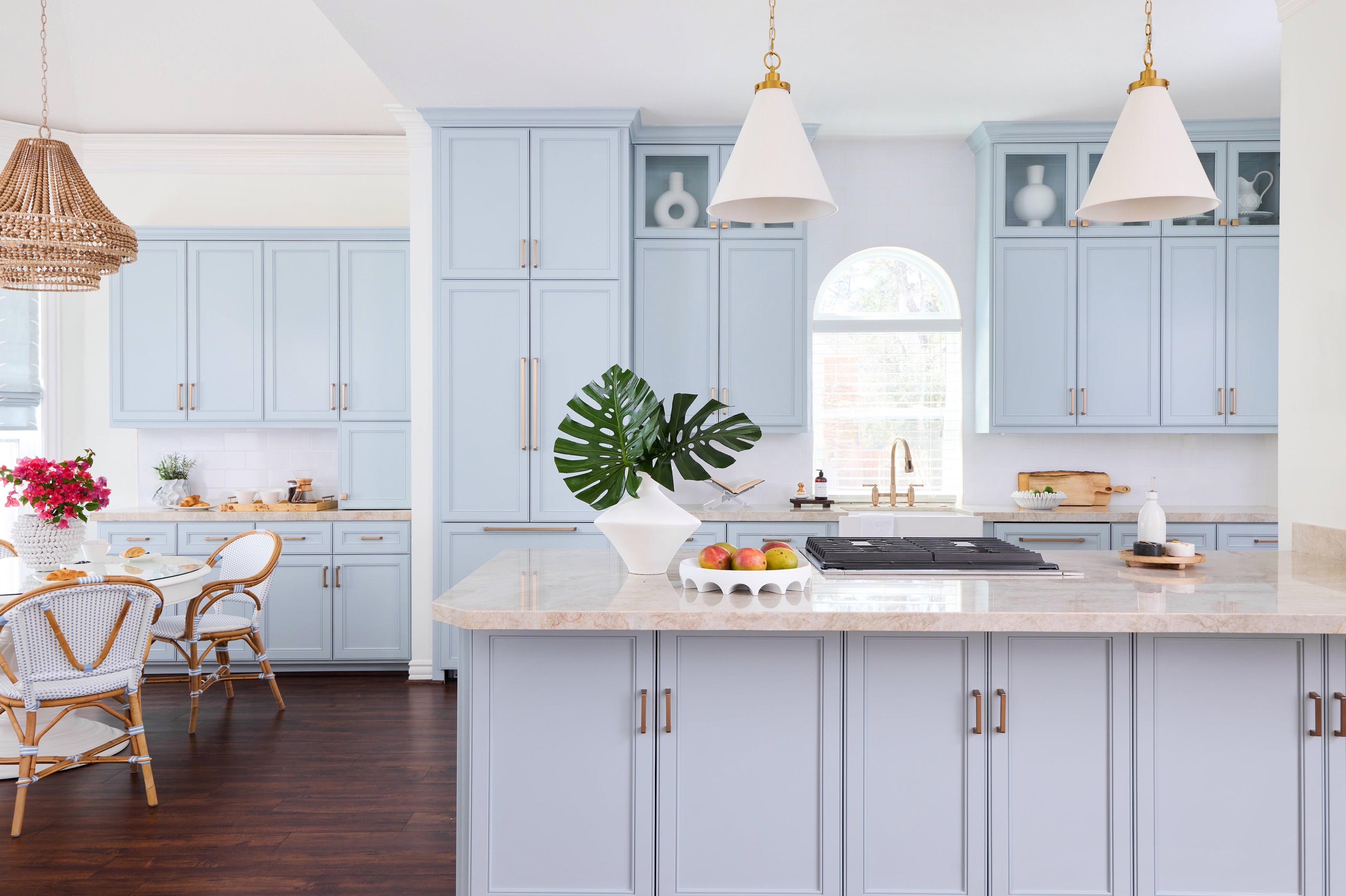Coastal kitchen with light blue cabinets, Taj Majal quartzite, hanging white and brass pendants over cooktop. White breakfast table to the left with white rattan chairs and wood beaded pendant.